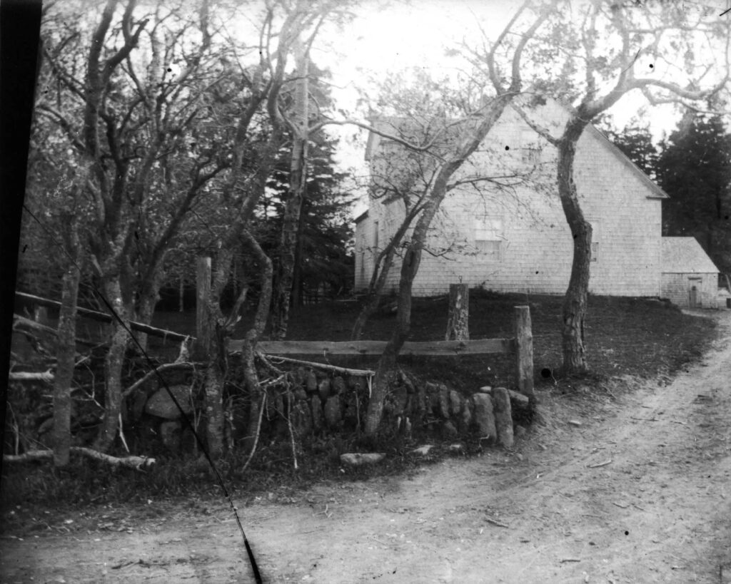 Image en noir et blanc d’une allée clôturée menant à une maison entourée d’arbres. Une petite fenêtre se trouve au centre à l’étage.