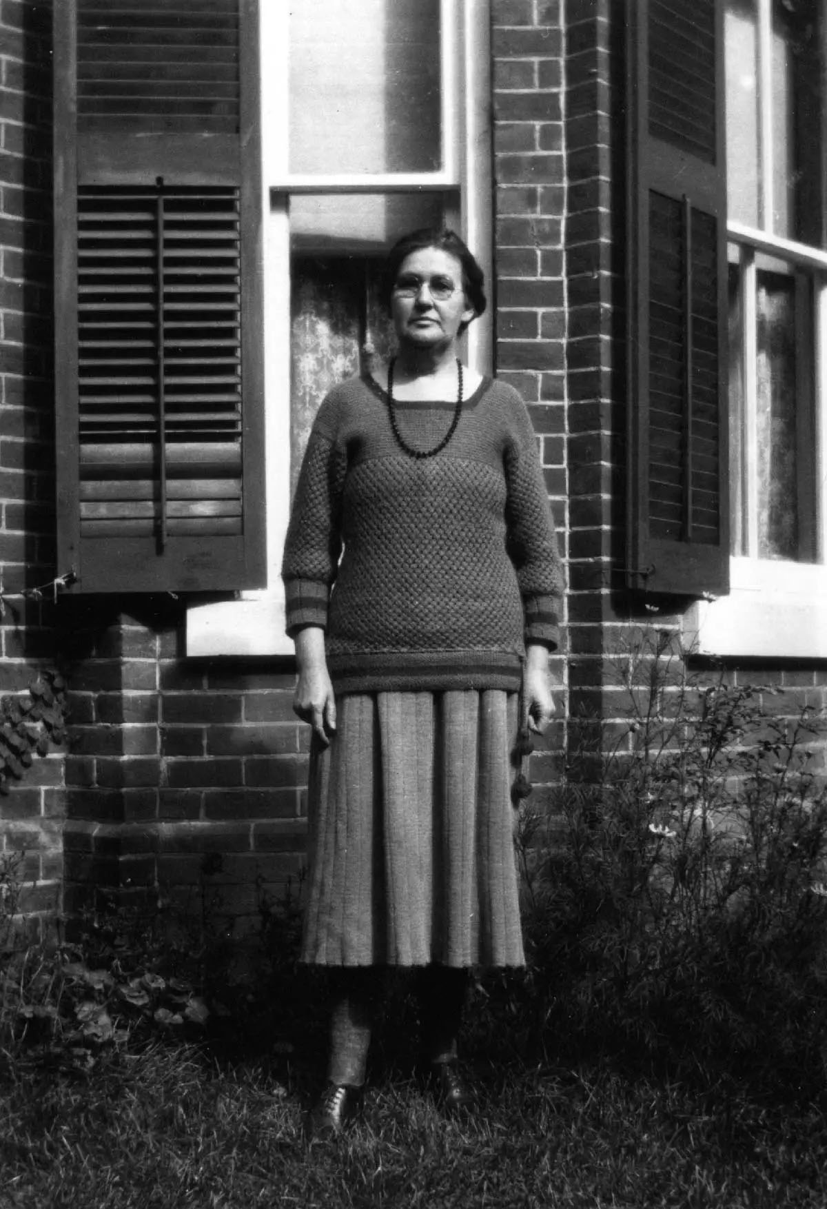 black and white image of a woman standing next to a brick home; she wears glasses and a long sweater with a skirt.