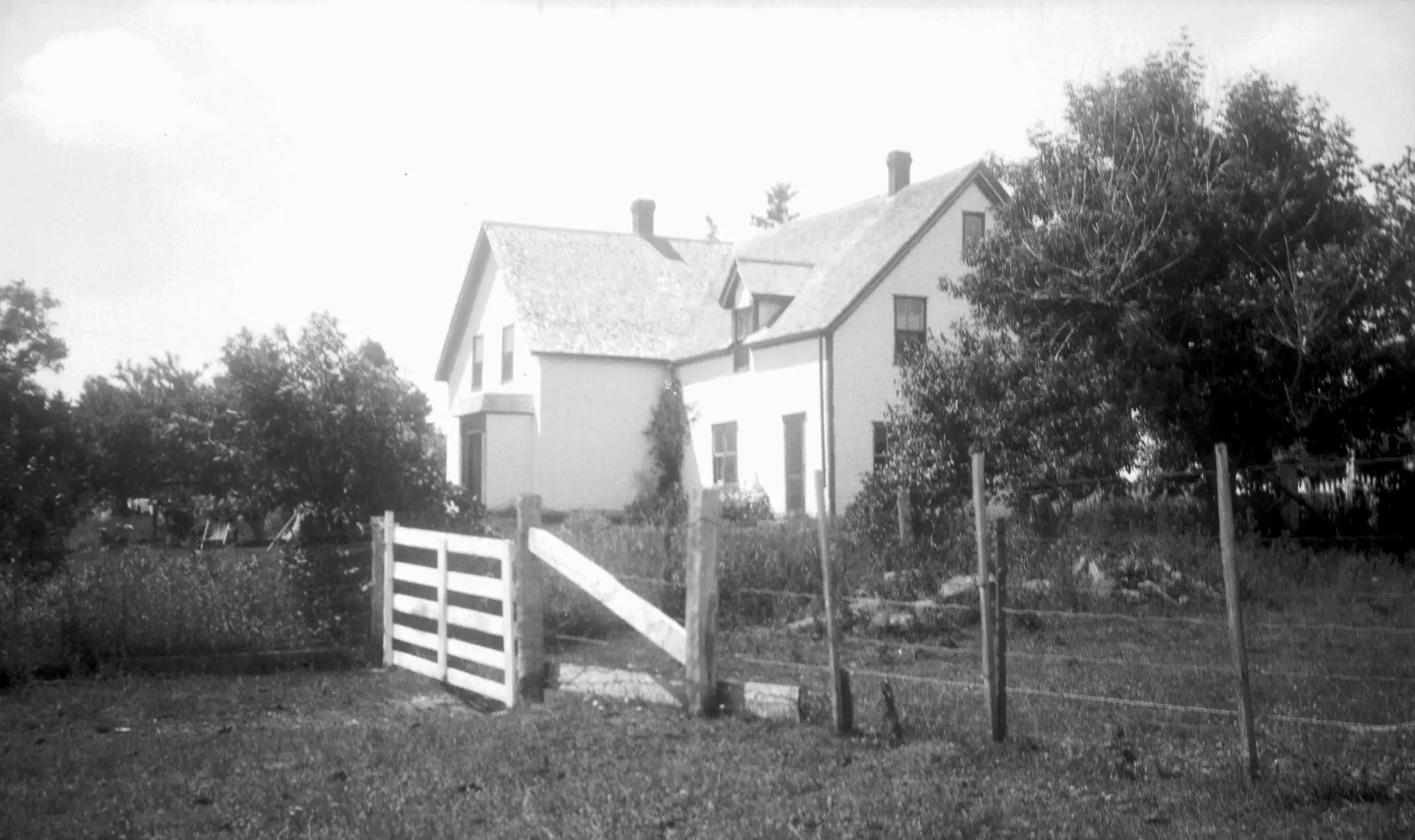 Une maison blanche à ossature en bois en forme de L, munie de trois cheminées, est entourée d’arbres; une clôture et une porte sont visibles au premier plan.