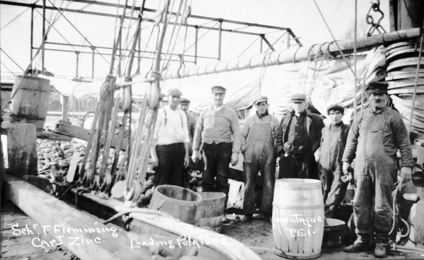 Seven men stand on the deck of a docked schooner, sails furled, with heaps of potatoes and some barrels visible.