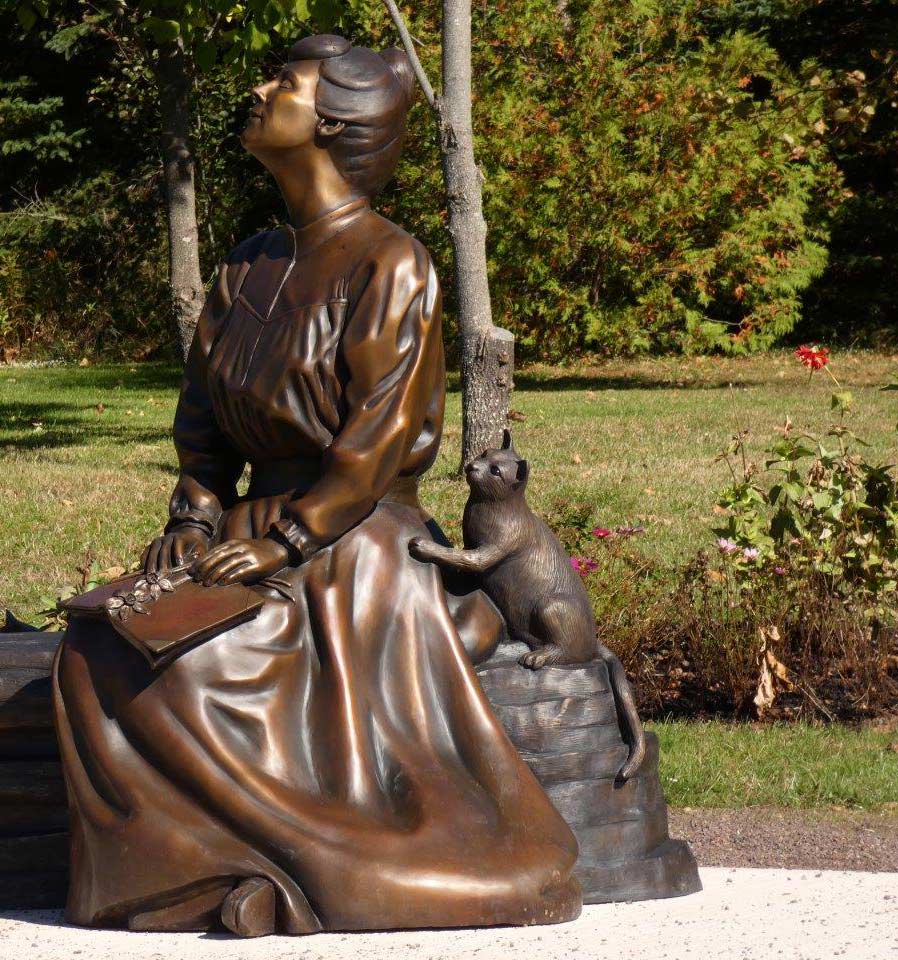 Statue en bronze d’une femme vêtue d’une jupe longue, un livre sur les genoux, le visage tourné vers le soleil.