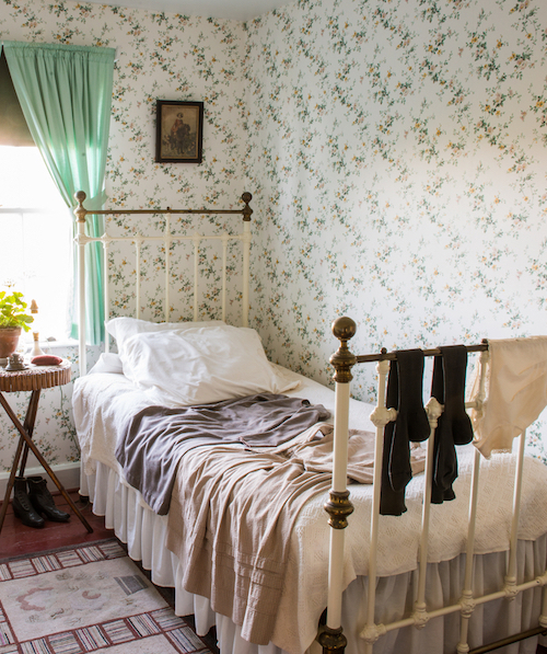 the corner of a sunny bedroom featuring a brass bed, dainty floral wallpaper and light curtains on the window