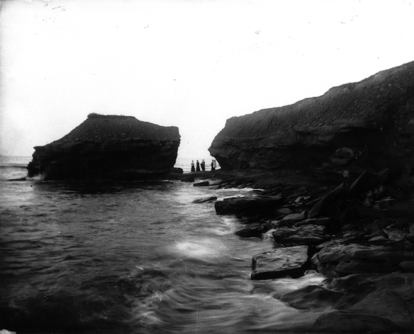 image en noir et blanc, à fort contraste, d'un rivage rocheux, avec de grandes formations rocheuses et de petits personnages au loin