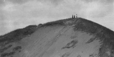 Photo d’une dune abrupte avec des herbes qui poussent tout en haut. On aperçoit trois silhouettes au sommet.