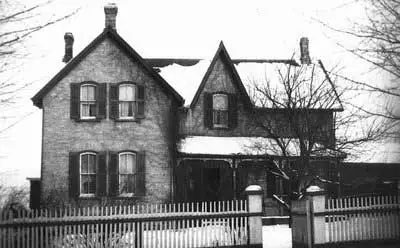 Photographie d’une maison en briques avec un pignon avant sur la gauche et un porche près du sol sur la droite.