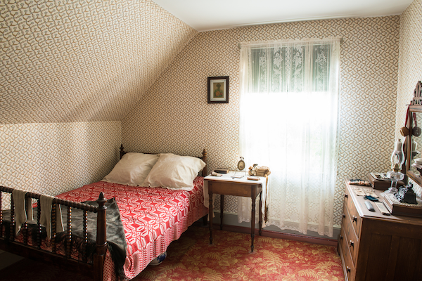 image d'une chambre à coucher ensoleillée avec une courtepointe rouge sur le lit, des rideaux en dentelle et une commode bien rangée à droite