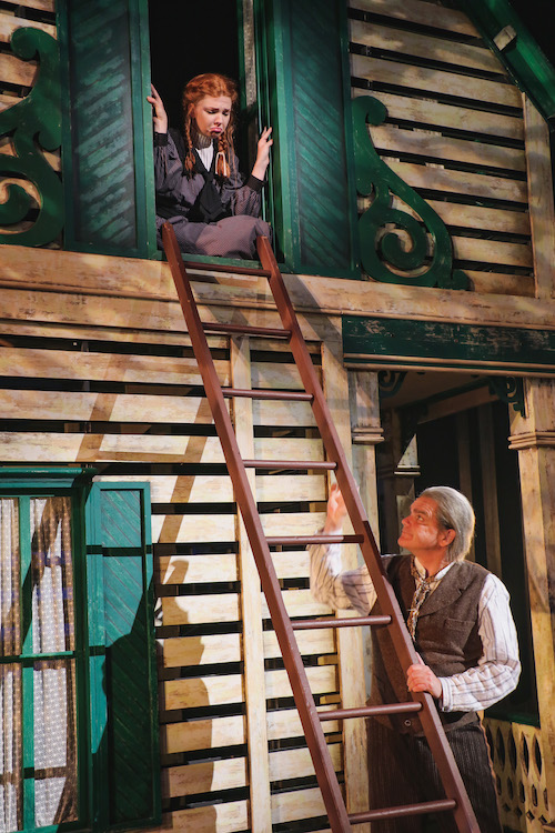 the facade of a white and green house on a stage, young girl in the upper window, and an older man about to ascend a ladder up to her.