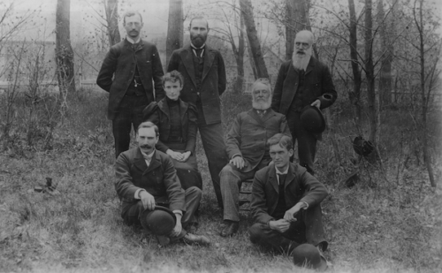six men and one woman pose in a small group outside