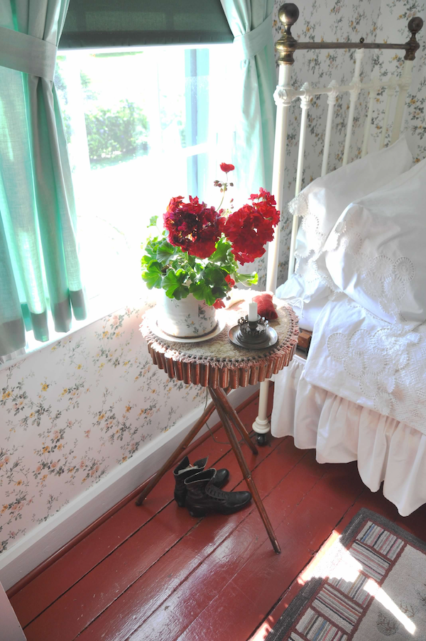 a sunny bedroom window with a small three-legged table and a geranium
