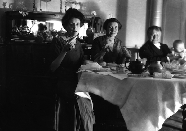 photo en noir et blanc de deux femmes et d’un homme tenant un bébé, souriant devant une table dressée pour le thé